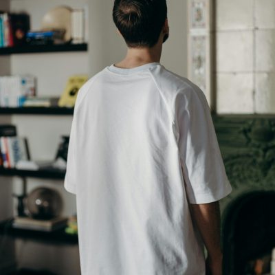 Adult male wearing a white t-shirt stands indoors facing a bookshelf.